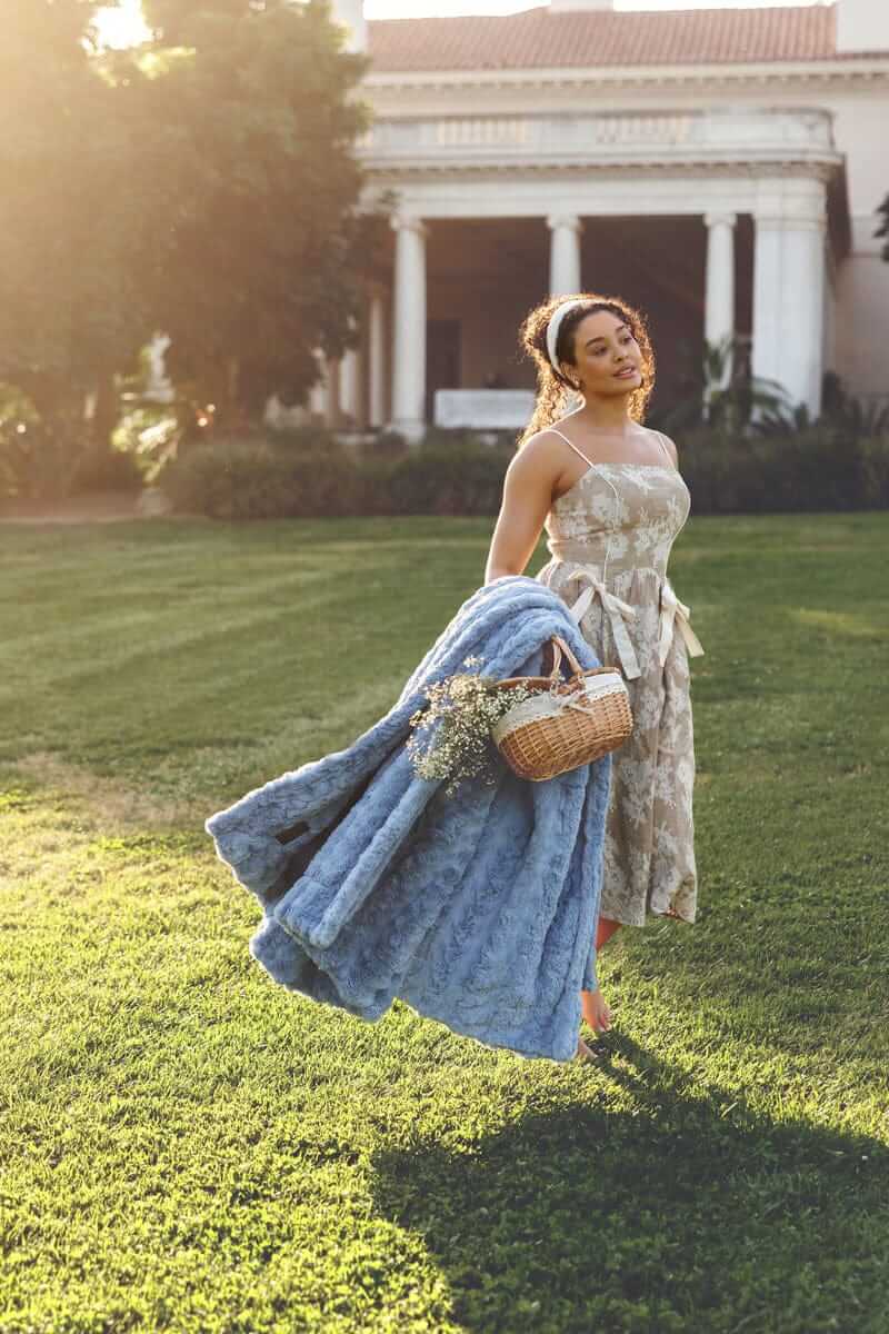 Woman holding a basket and an Ultra Sorbet Faded Denim Blanket while standing in a sunlit garden.