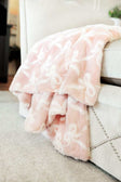 Bows Blush Blanket draped over a stylish chair, showcasing its soft blush pink color and embossed bow pattern.