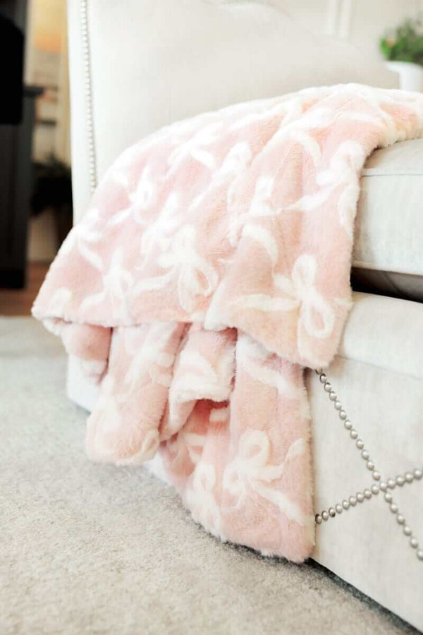 Bows Blush Blanket draped over a stylish chair, showcasing its soft blush pink color and embossed bow pattern.