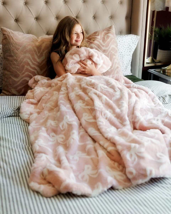 A girl snuggles under the cozy Bows Blush Blanket featuring a soft pink color and embossed bow pattern.