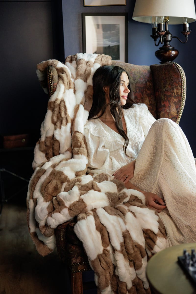 A cozy scene featuring a woman draped in the Hugs Check Milk Chocolate Blanket on a luxurious armchair.