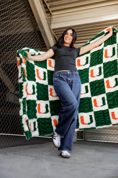 A happy woman showing off a Miami Hurricanes Blanket with a green and white checkered pattern.