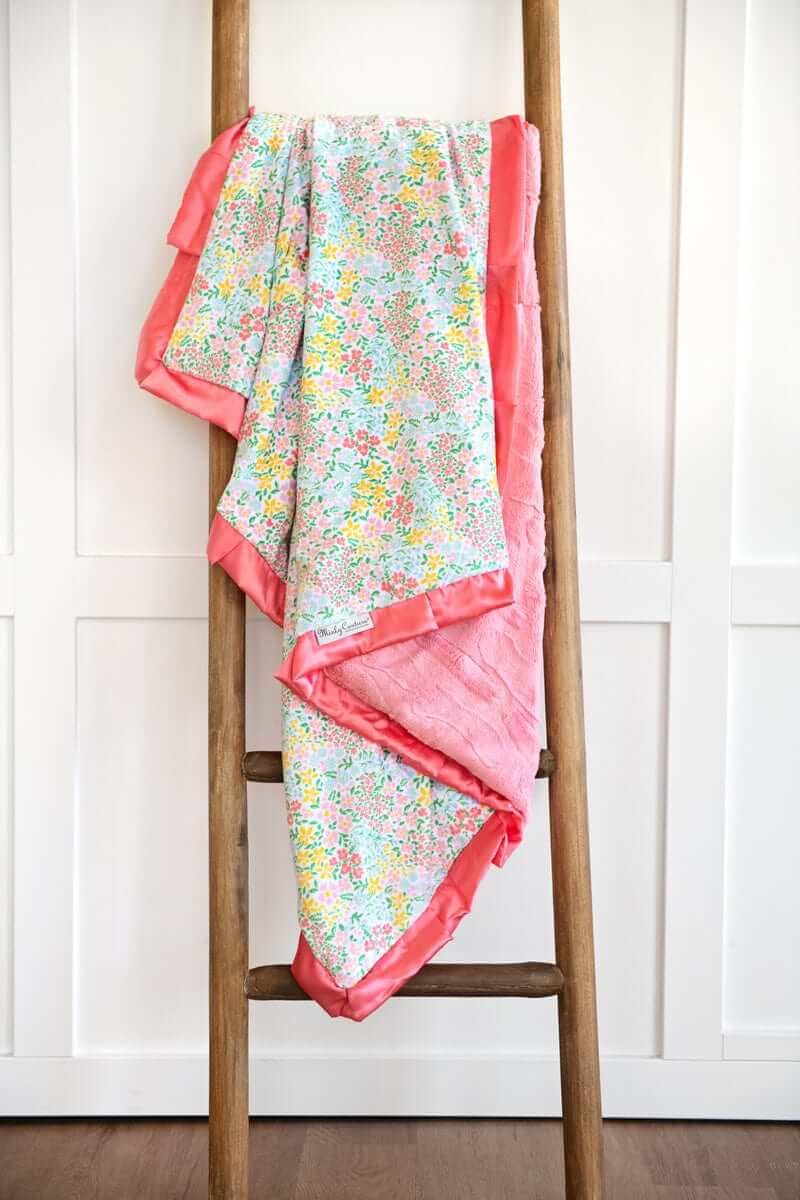 Dainty Floral Pastel Blanket displayed on a wooden ladder, featuring small colorful flowers and a coral satin ruffle.