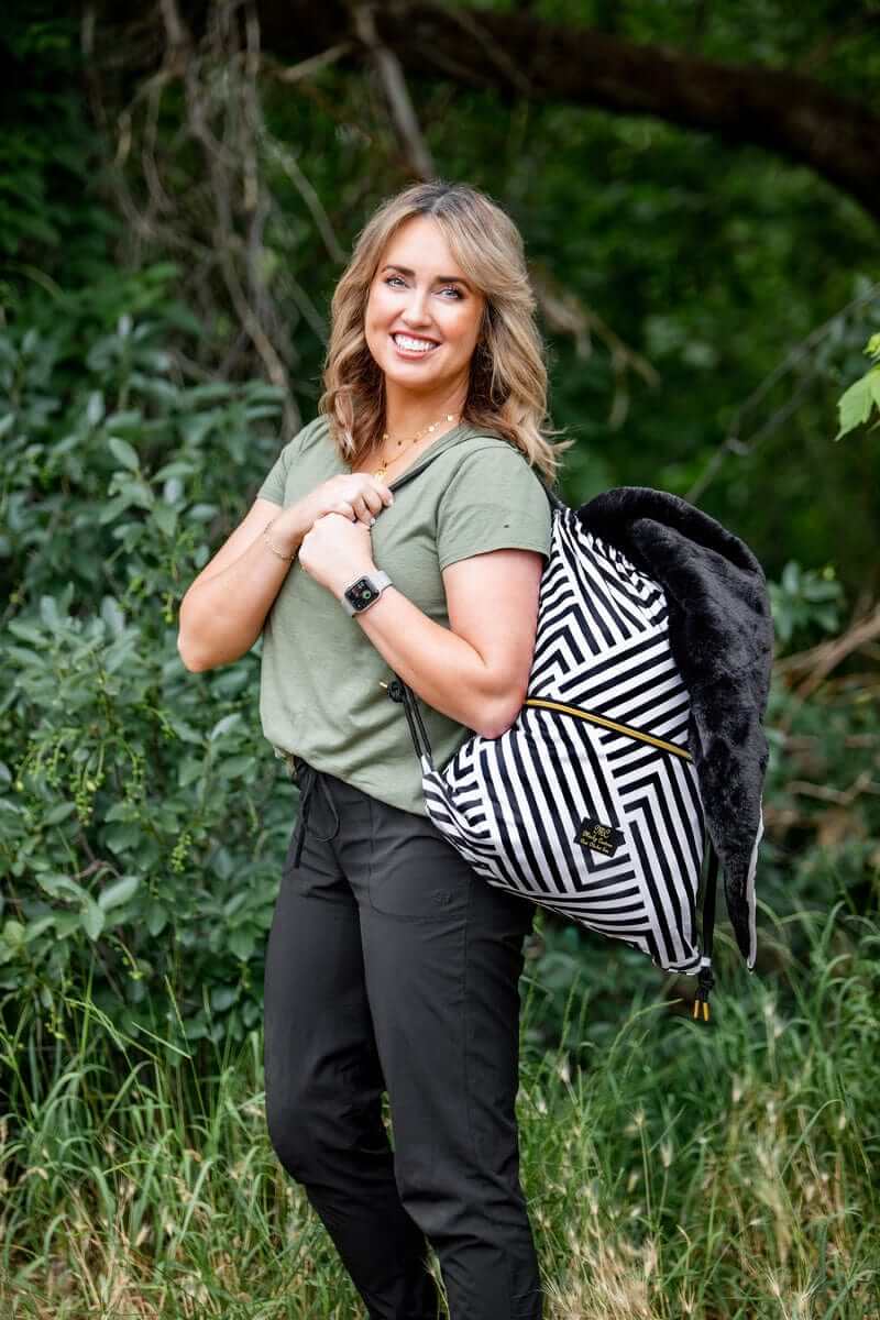 Woman smiling outdoors with Black Geo Everywhere Blanket backpack in a striped pattern against greenery.
