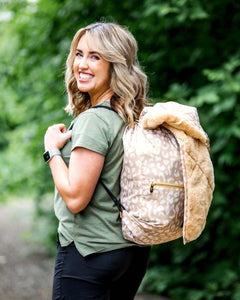 Everywhere Blanket Tan Cheetah styled as a backpack on a woman smiling outdoors, perfect for picnics and camping.