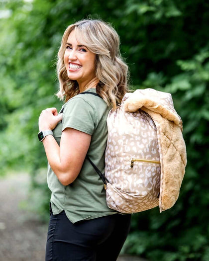 Everywhere Blanket Tan Cheetah styled as a backpack on a woman smiling outdoors, perfect for picnics and camping.