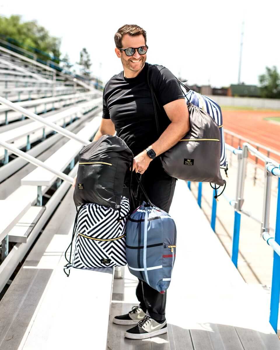 Man carrying multiple stylish bags while walking on stadium bleachers wearing sunglasses and athletic clothing.