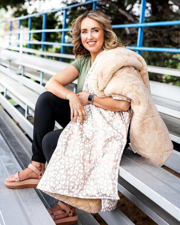 Woman sitting on bleachers with Tan Cheetah Everywhere Blanket, cozy and stylish for outdoor events.