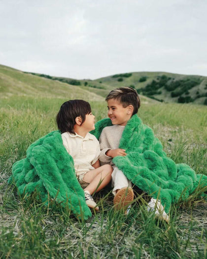 Two children sitting on grass, wrapped in an Ultra Sorbet Kelly Green weighted blanket, enjoying a cozy moment outdoors.