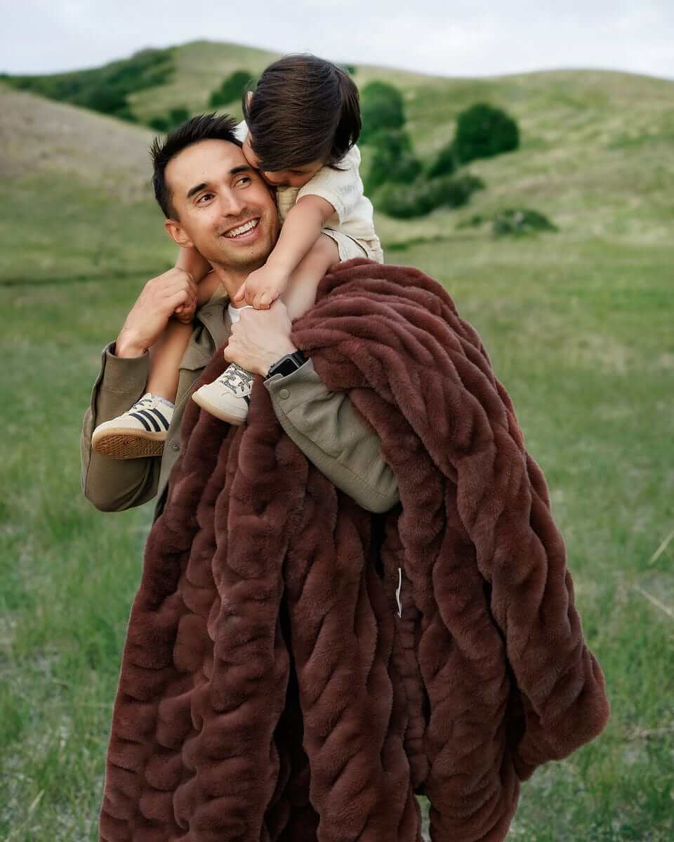 A person carrying a child on their shoulders while wrapped in a rich mahogany colored blanket, standing in an open field.
