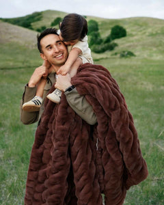 A person carrying a child on their shoulders while wrapped in a rich mahogany colored blanket, standing in an open field.