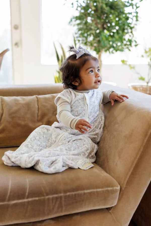 Baby girl sitting on a light brown couch, with a Forget Me Not Wearable Minky Blanket.