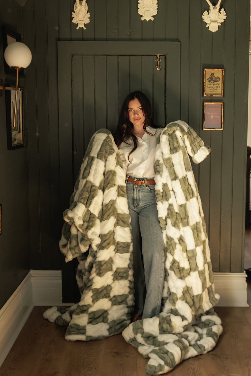 Hugs Check Sage Blanket displayed by a model in a stylish interior, showcasing its soft checkered design.