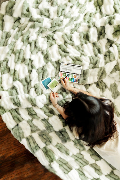 A person relaxing on the Hugs Check Sage Blanket while reviewing art materials and enjoying a cozy atmosphere.