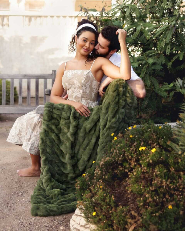 Cozy couple wrapped in the Hugs Camp Green Blanket sitting on a bench surrounded by greenery.