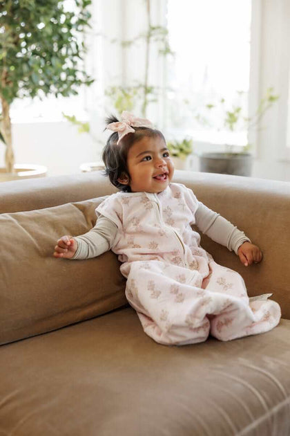 Smiling baby girl sitting on a light brown couch, with a Hampton Blooms Wearable Minky Blanket.