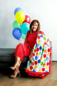 Woman in red dress holding Happy Birthday Blanket with colorful balloon print and vibrant balloons in the background.