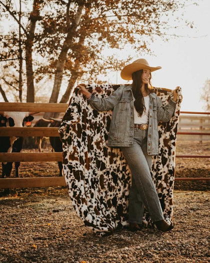 Model showcasing the Hugs Brown Cow faux fur blanket in a rustic setting, surrounded by cows and fall foliage.