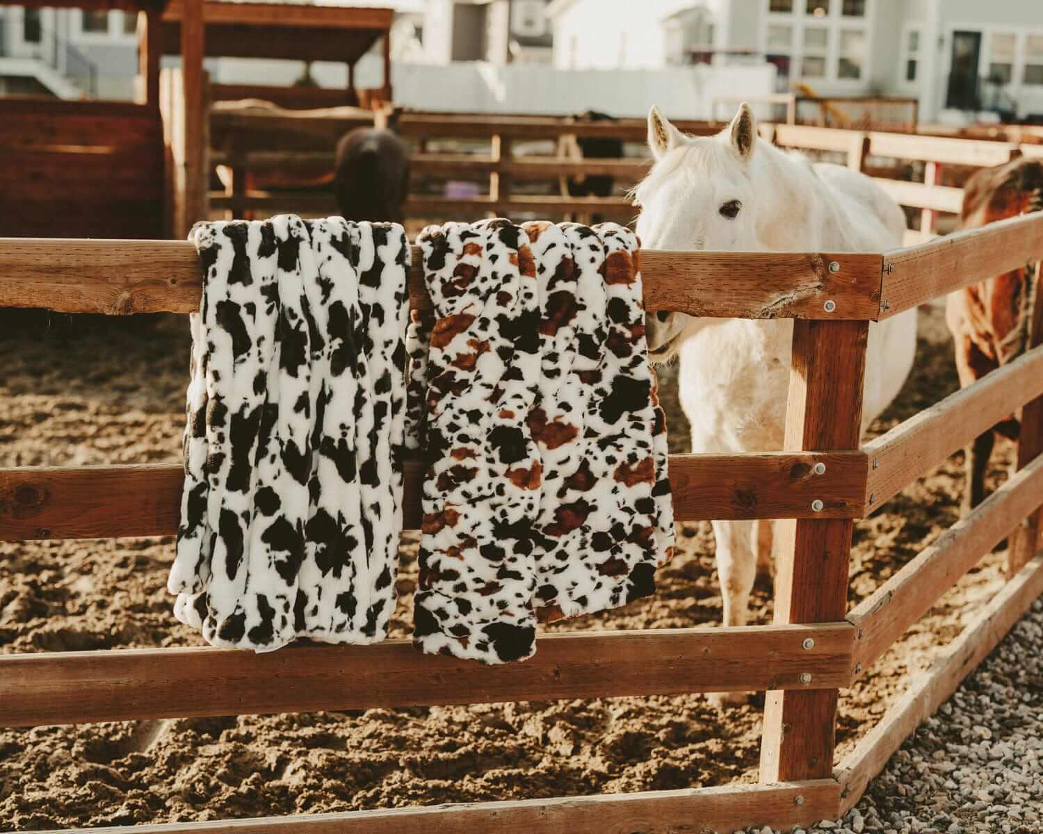 Hugs Brown Cow faux fur blanket displayed on a fence next to a horse in a barn, showcasing its soft cow print design.