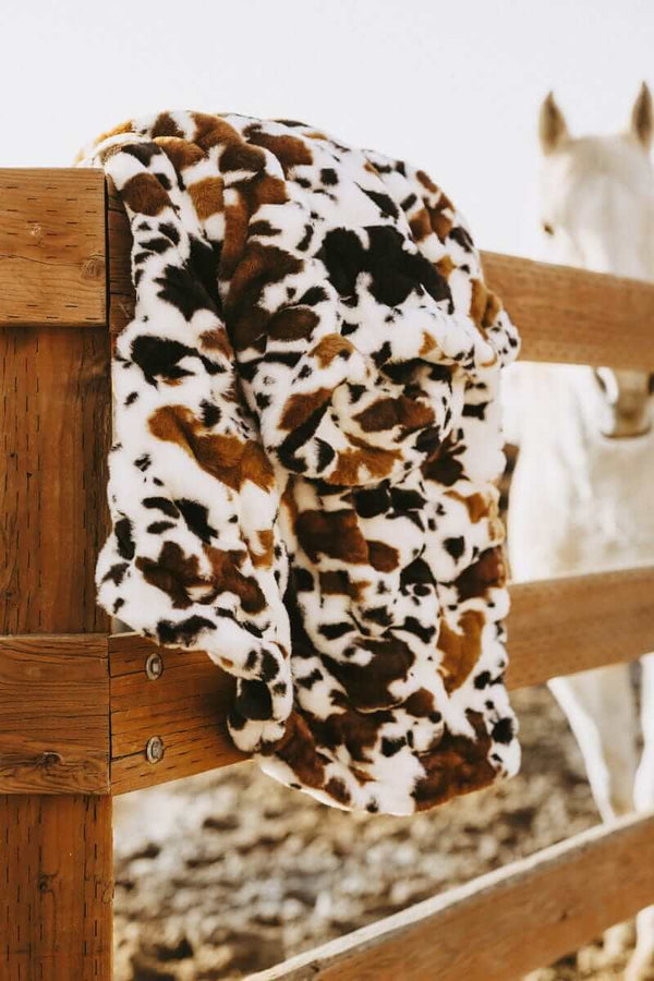 Hugs Brown Cow Blanket draped over a wooden fence with a horse in the background, featuring a soft cow print design.