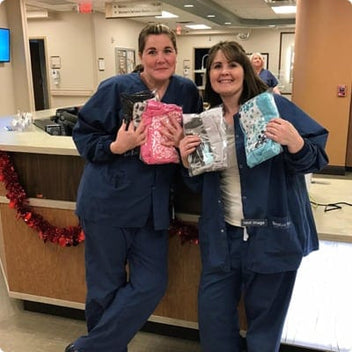 Two healthcare professionals in scrubs smiling while holding colorful fabric bundles in a hospital setting.