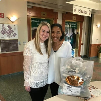Two women smiling together at an event, one holding a gift bag, in a well-lit lobby area.