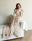 A woman wrapped in the Hugs Topaz faux fur Blanket, enjoying its soft warmth on a couch.