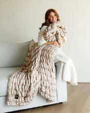 A woman wrapped in the Hugs Topaz faux fur Blanket, enjoying its soft warmth on a couch.