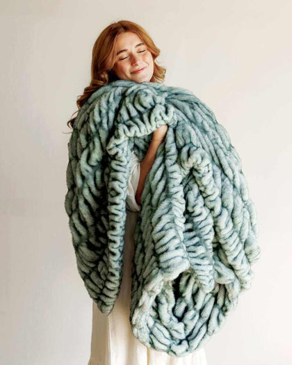 Hugs Jade Blanket wrapped around a woman, showcasing its plush, jade color and luxurious faux fur design.
