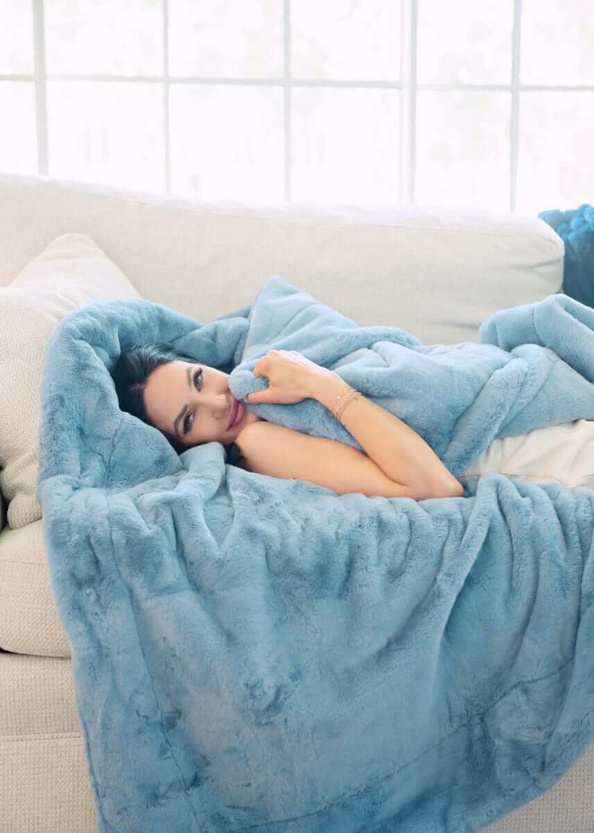 Model relaxing under a Lush Denim Blanket on a couch, showcasing its fluffy blue fabric and cozy comfort.