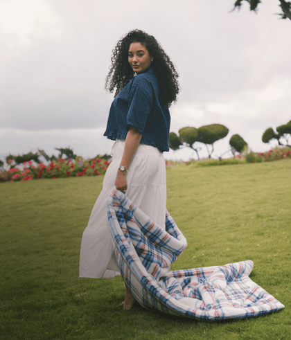 Woman with curly hair in blue shirt holding a plaid blanket, standing on green grass with flowers in the background.