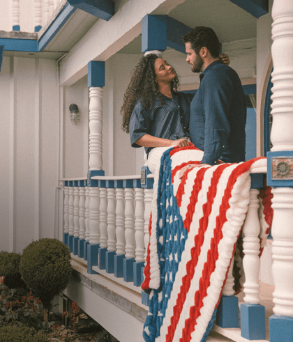 Couple sharing a romantic moment on a porch, wrapped in an American flag blanket, surrounded by greenery.