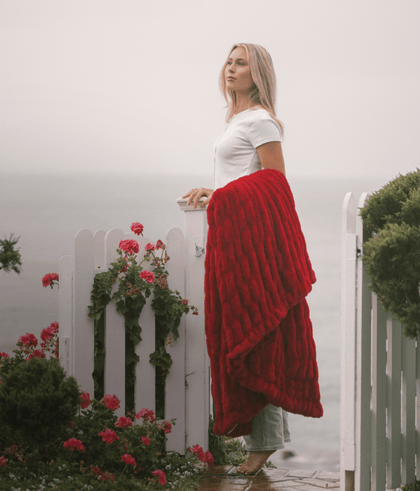 Woman in a white top draped with a red blanket, standing by a white fence and ocean view, surrounded by flowers.