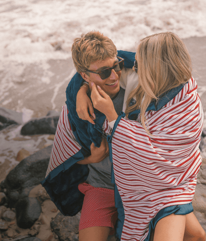 Young couple joyfully embracing on a beach, wrapped in a striped blanket, enjoying a sunny day by the waves.