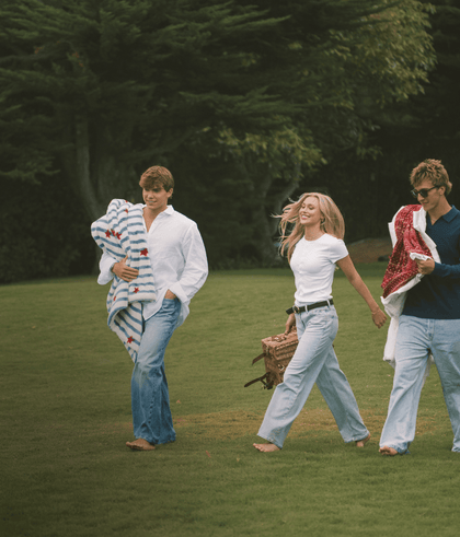 Three friends walking on a grassy field, casually dressed, carrying blankets and enjoying a day outdoors.