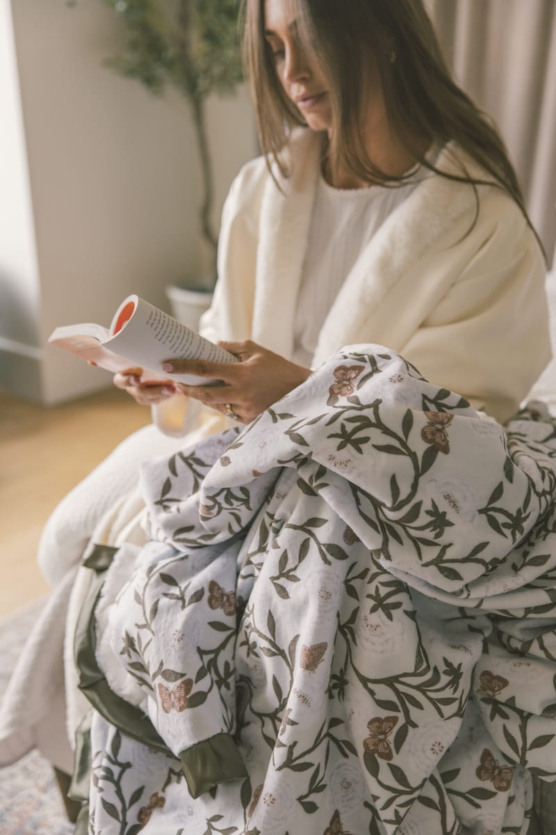 Woman sitting on a couch with a floral blanket, holding a magazine.