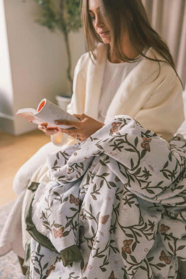 Woman sitting on a couch with a floral blanket, holding a magazine.