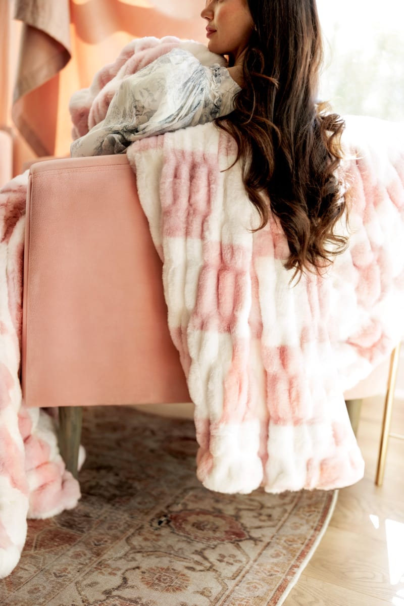A woman lounges on a pink chair wrapped in the Hugs Check Pink Blanket featuring a soft white and light pink checkered pattern.