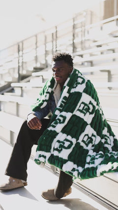 Man sitting outdoors wrapped in a green and white Minky Couture blanket with a bold logo pattern.
