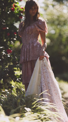 Woman in a floral dress holding a Minky Easter Floral Blanket surrounded by greenery in a sunlit garden.