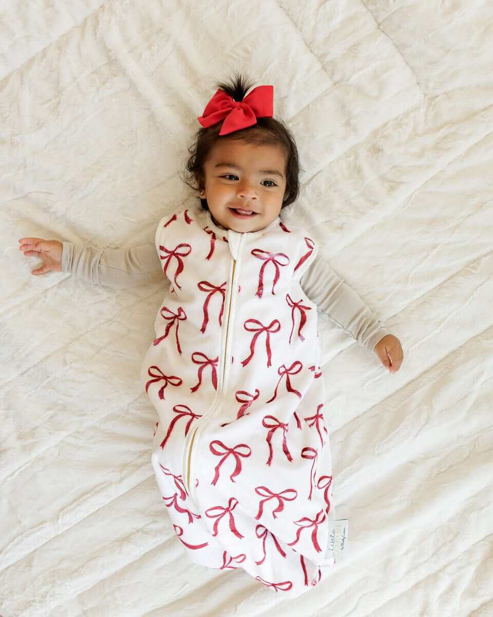 Smiling baby girl lying on a bed with Minky Couture red ribbon wearable blanket.