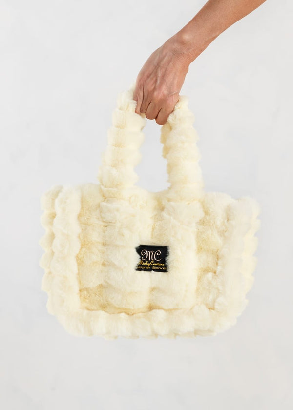A butter yellow colored, fluffy faux-fur handbag with double shoulder straps, held by a person's hand against a white background. A black "MC" Minky Couture brand patch with gold lettering is attached to the front.