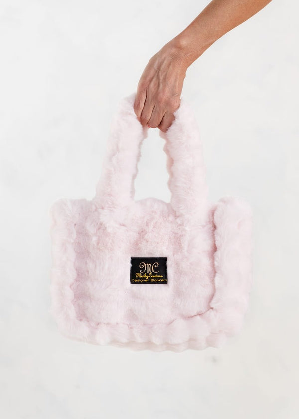 A light pink fluffy faux-fur handbag held by a person's hand against a clean white background. A black "MC" Minky Couture logo patch with gold lettering is attached to the front.