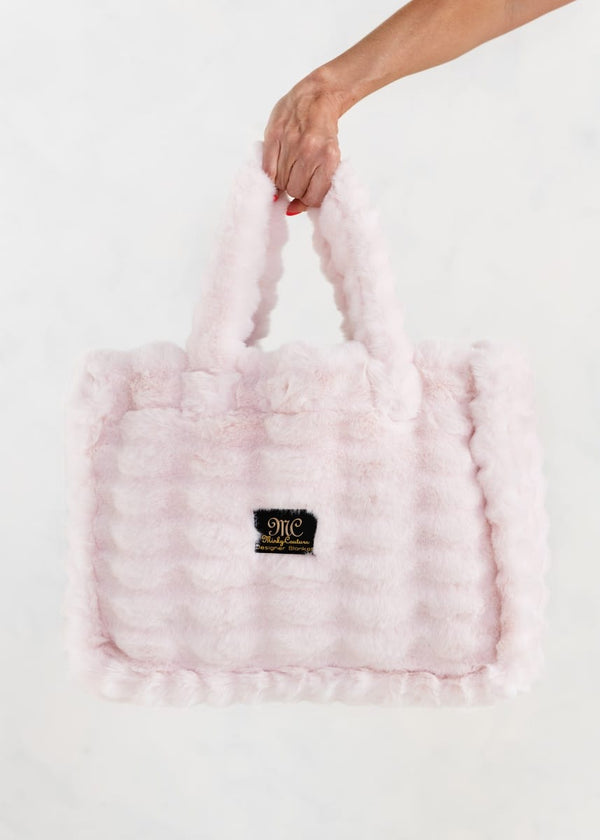 A light pink fluffy faux-fur bag with a textured wavy pattern, held by a hand against a plain white background. A small black "MC" Minky Couture logo patch with gold lettering is visible on the front.