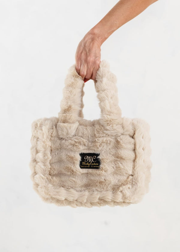 A hand holding a small fluffy faux-fur tote bag in a beige color, featuring a black rectangular logo patch on the front with gold lettering, set against a plain white background.