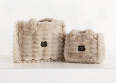 Two fluffy faux-fur handbags in a light beige color, one larger bag and one smaller bag, both featuring a black and gold 'MC' Minky Couture logo patch on the front, displayed on a white surface against a white background.