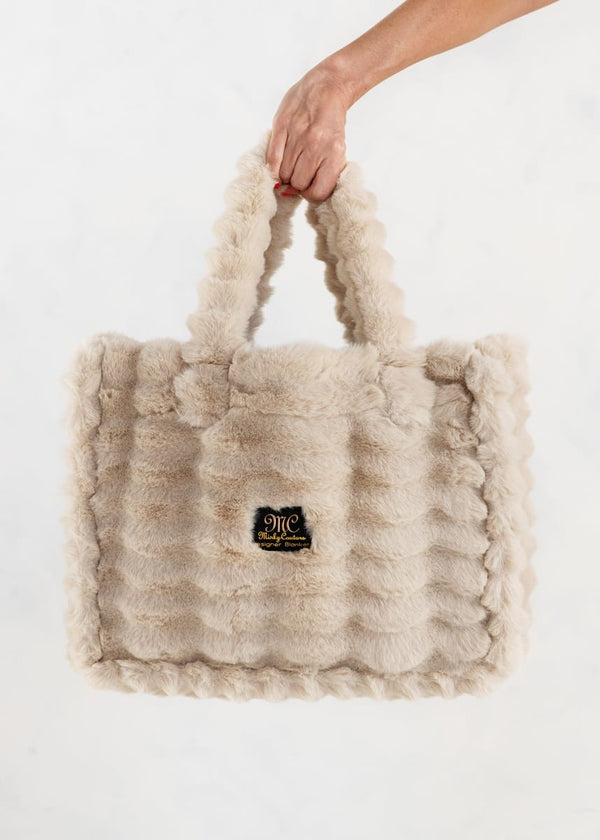 A hand holding a fluffy, textured beige shoulder bag with a small black and gold logo patch on the front, set against a plain white background.