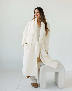 Woman wearing a cozy Cream Robe with plush lining, standing beside a soft stool.