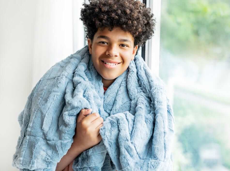 A smiling child wrapped in a soft, muted blue Sorbet Denim Blanket, showcasing its cozy texture.