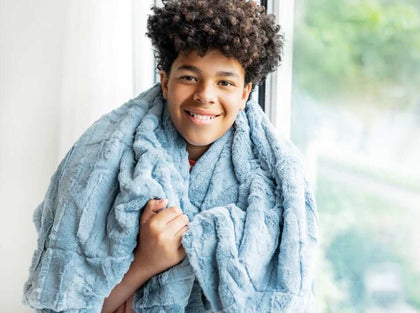 A smiling child wrapped in a soft, muted blue Sorbet Denim Blanket, showcasing its cozy texture.